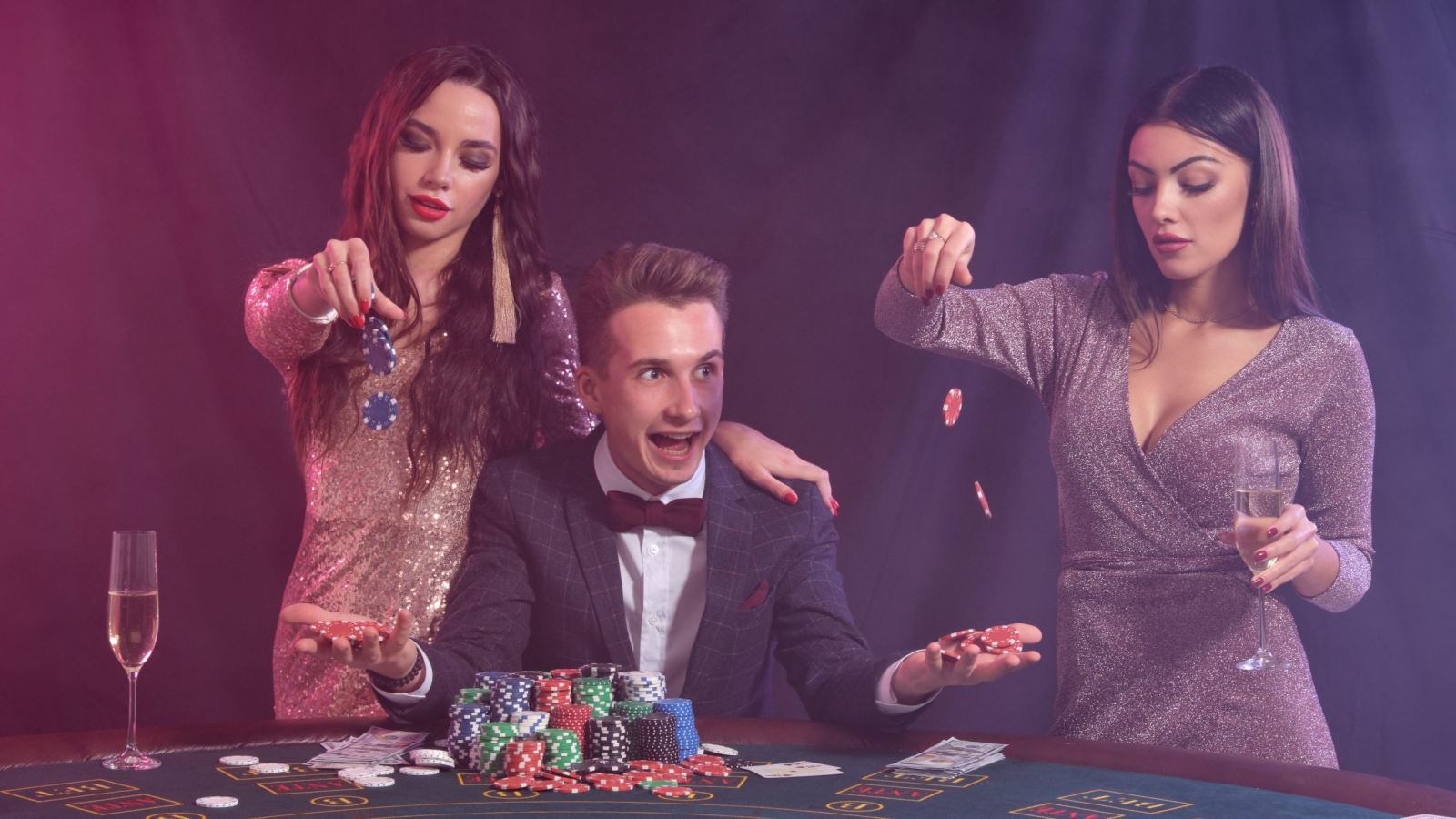 Man playing poker at casino sitting at table with stacks of chips, money, cards. Celebrating win with two brunette women. Black, smoke background, colorful backlights. Gambling, alcohol. Close-up.