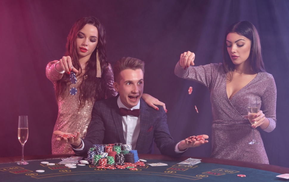 Man playing poker at casino sitting at table with stacks of chips, money, cards. Celebrating win with two brunette women. Black, smoke background, colorful backlights. Gambling, alcohol. Close-up.